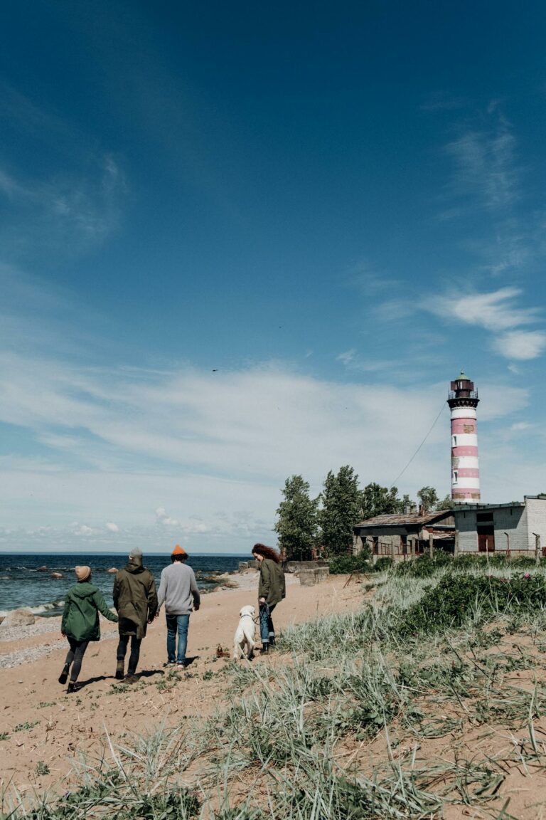 Family and dog walking along sandy beach by a lighthouse on a clear summer day.